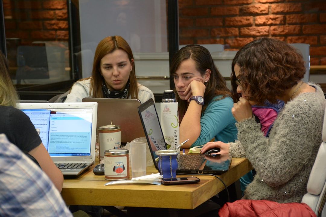 Three colleagues collaborating on laptops at a shared workspace with a brick wall background.
